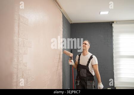 A man is using a paint roller to apply paint to a wall. He is dressed in work clothes and protective gear. The wall is a blank canvas being transforme Stock Photo