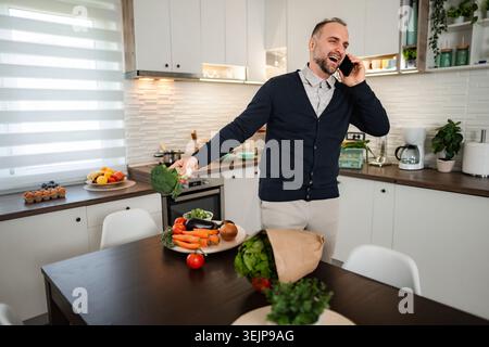 Man laughing talking on smartphone holding broccoli in kitchen Stock Photo