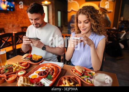 Happy couple taking photos of their food using smartphones, sitting at a table in a restaurant Stock Photo