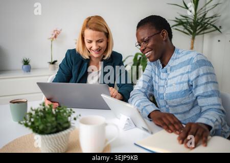 Diverse women collaborating on laptop and smiling at work Stock Photo