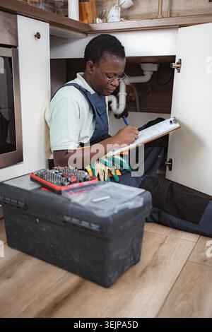 Female plumber checking pipes and writing on clipboard in kitchen Stock Photo