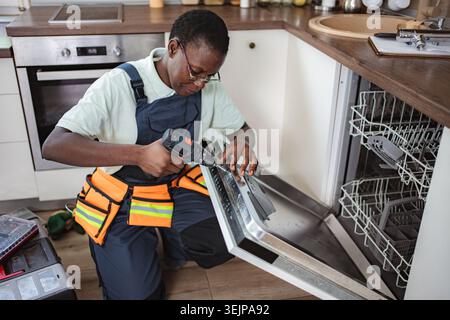 Technician repairing dishwasher using electric screwdriver in kitchen Stock Photo