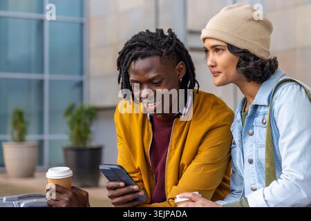 Diverse friends sitting outside office, woman in beanie leaning, sharing phone and to-go cups. Outdoors, urban, campus, casual, Stock Photo
