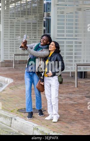 Diverse couple wearing jackets and scarves taking phone selfie holding skateboard on brick sidewalk. Duo, urban, streetstyle, outdoor, casual, sociabl Stock Photo