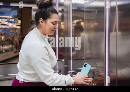 Woman pressing elevator button while using smartphone in a mall Stock Photo