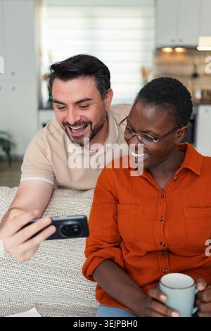 Happy multi-ethnic couple laughing while watching videos on smartphone at home Stock Photo