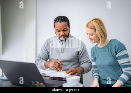 Diverse couple discussing documents and working on laptop at home Stock Photo