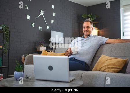 Middle aged Caucasian man sitting on the sofa watching videos on laptop Stock Photo