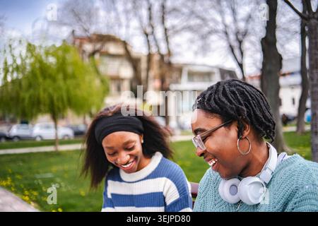 Two young adult women friends laughing outdoors together on a park bench Stock Photo