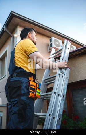 Electrician with tool belt climbing a ladder and installing an air conditioner on a building facade Stock Photo