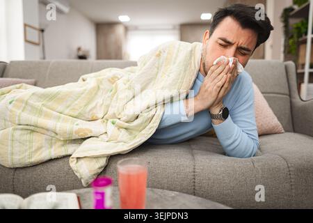 Sick man having a runny nose while lying on a sofa covered with blanket Stock Photo