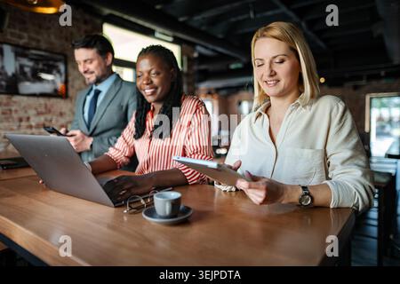 Three diverse business professionals working, communicating using digital devices in a modern cafe Stock Photo