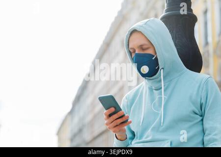 Man on street wears respiratory mask, walks through city, uses modern mobile phone concentrated into display uses wireless inter Stock Photo