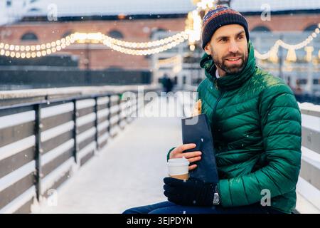 Middle aged man looks thoughtfully aside, has snack outdoors during winter weather, drinks hot coffee and eats roll, walks on st Stock Photo