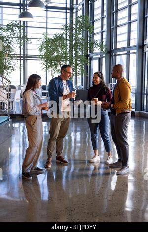 Diverse coworkers in business attire standing and discussing in atrium with coffee cups, tablet. Office, collaboration, colleagues, discussion, plants Stock Photo