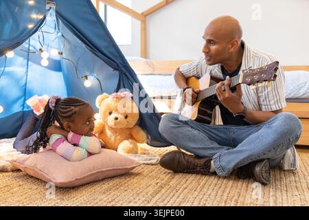 African American father and daughter sitting on rug, father playing acoustic guitar by blue teepee. Cozy, music, home, bonding, family, warm, natural Stock Photo