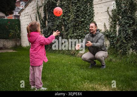 Father and daughter playing catch with orange ball in backyard Stock Photo