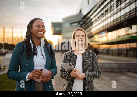 Two diverse businesswomen are smiling and walking together in a city at sunset Stock Photo