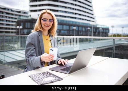 Businesswoman working remotely outdoors, using smartphone and laptop Stock Photo