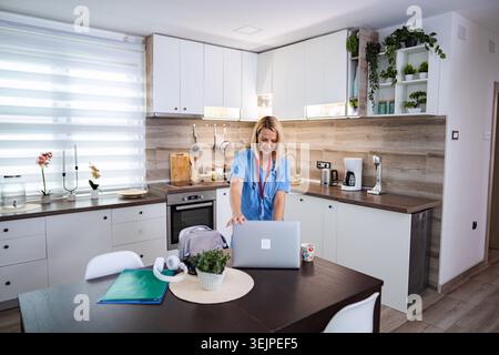 Female Doctor Wearing Blue Scrubs is Opening Her Laptop in the Kitchen Stock Photo