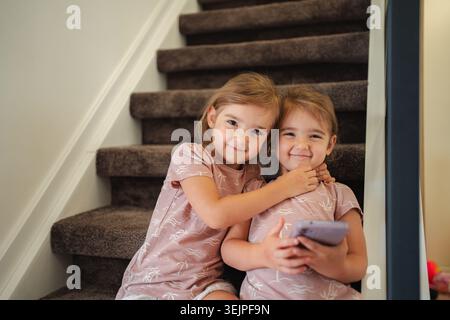 Two preschool age twin sisters hugging and using smartphone on stairs at home Stock Photo
