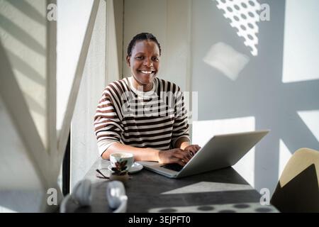 Smiling woman using laptop sitting at cafe by window Stock Photo
