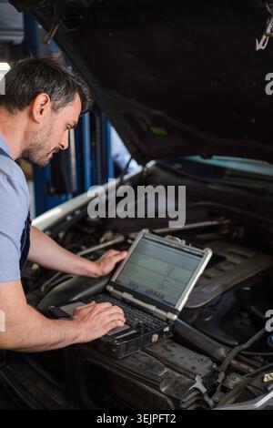 Auto mechanic performing diagnostics on a car engine using a laptop in a repair shop Stock Photo