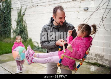 Father pushing daughter on swing with another daughter playing Stock Photo