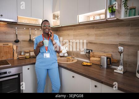 Smiling african american female doctor drinking coffee and using smartphone in kitchen Stock Photo