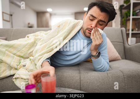 Sick man having a runny nose while lying on a sofa covered with blanket Stock Photo