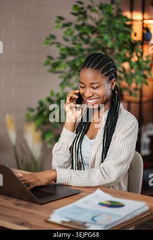 Black woman smiling talking on phone and typing laptop Stock Photo