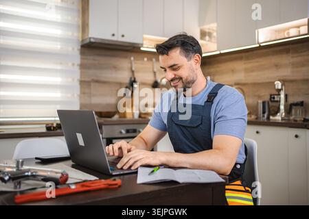 Smiling repairman working on laptop in kitchen with plumbing tools, using computer for online learning Stock Photo