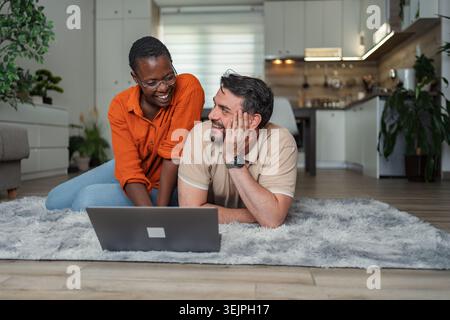 Happy multi-ethnic couple lying on carpet using laptop at home Stock Photo