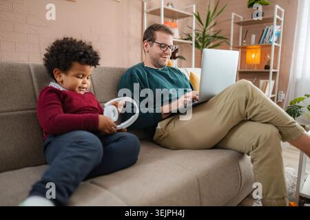 Father working on laptop at home next to his son Stock Photo