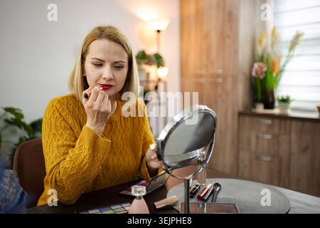 Young woman applying lipstick while seated at a stylish vanity in a cozy modern room with warm lighting Stock Photo
