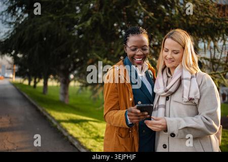 Diverse women friends smiling using smartphone outdoors Stock Photo