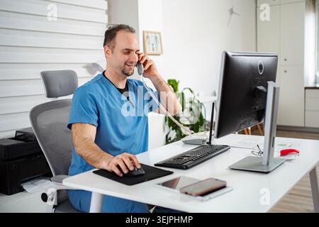 Smiling male doctor wearing scrubs talking on landline phone and using computer Stock Photo