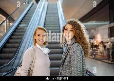 Two women friends smiling on escalator in shopping mall Stock Photo