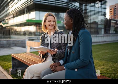 Two smiling businesswomen sitting on a bench, collaborating on a tablet outdoors Stock Photo