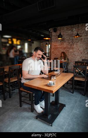 Freelancer working remotely from a cafe, talking on the phone and typing on his laptop Stock Photo