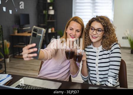 Two female colleagues are taking selfies and drinking coffee Stock Photo