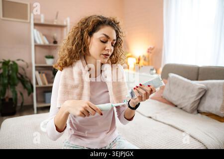 Woman applying toothpaste to electric toothbrush in bedroom Stock Photo