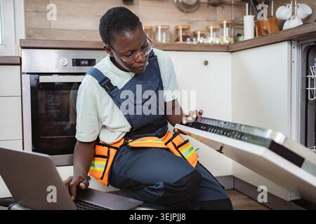 Repairwoman using laptop while repairing dishwasher in kitchen Stock Photo