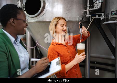 Female brewmasters working in craft brewery, tasting fresh beer from fermentation tank Stock Photo