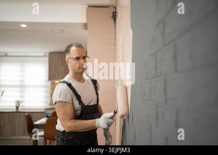 A man is using a paint roller to apply paint onto a wall. He is focused on the task at hand, ensuring even coverage and a smooth finish. The wall is b Stock Photo