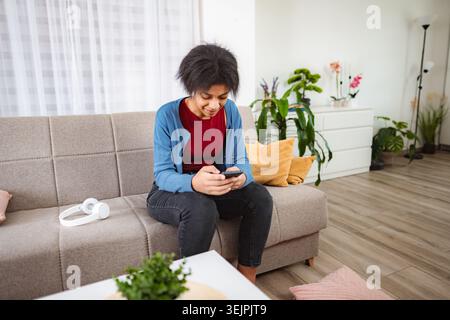Young woman using smartphone sitting on sofa in living room Stock Photo