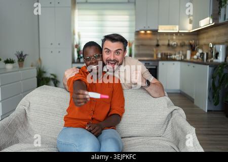 Excited multi-ethnic couple sharing pregnancy test result at home Stock Photo