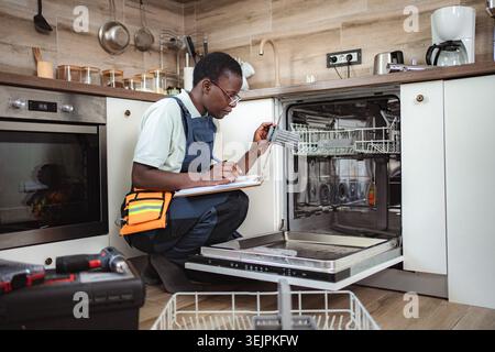 Technician inspecting dishwasher and writing on clipboard in kitchen Stock Photo