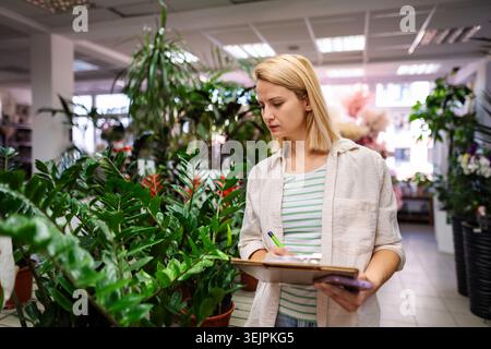 Female florist taking notes on clipboard while checking plants in flower shop Stock Photo