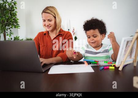 Mother working on laptop with child drawing at home Stock Photo